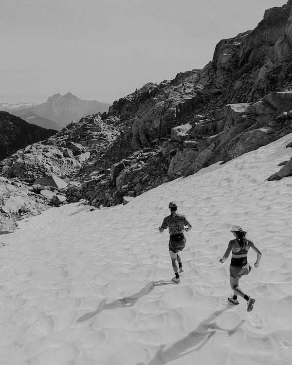 Two runners crossing a snowfield below rocky peaks